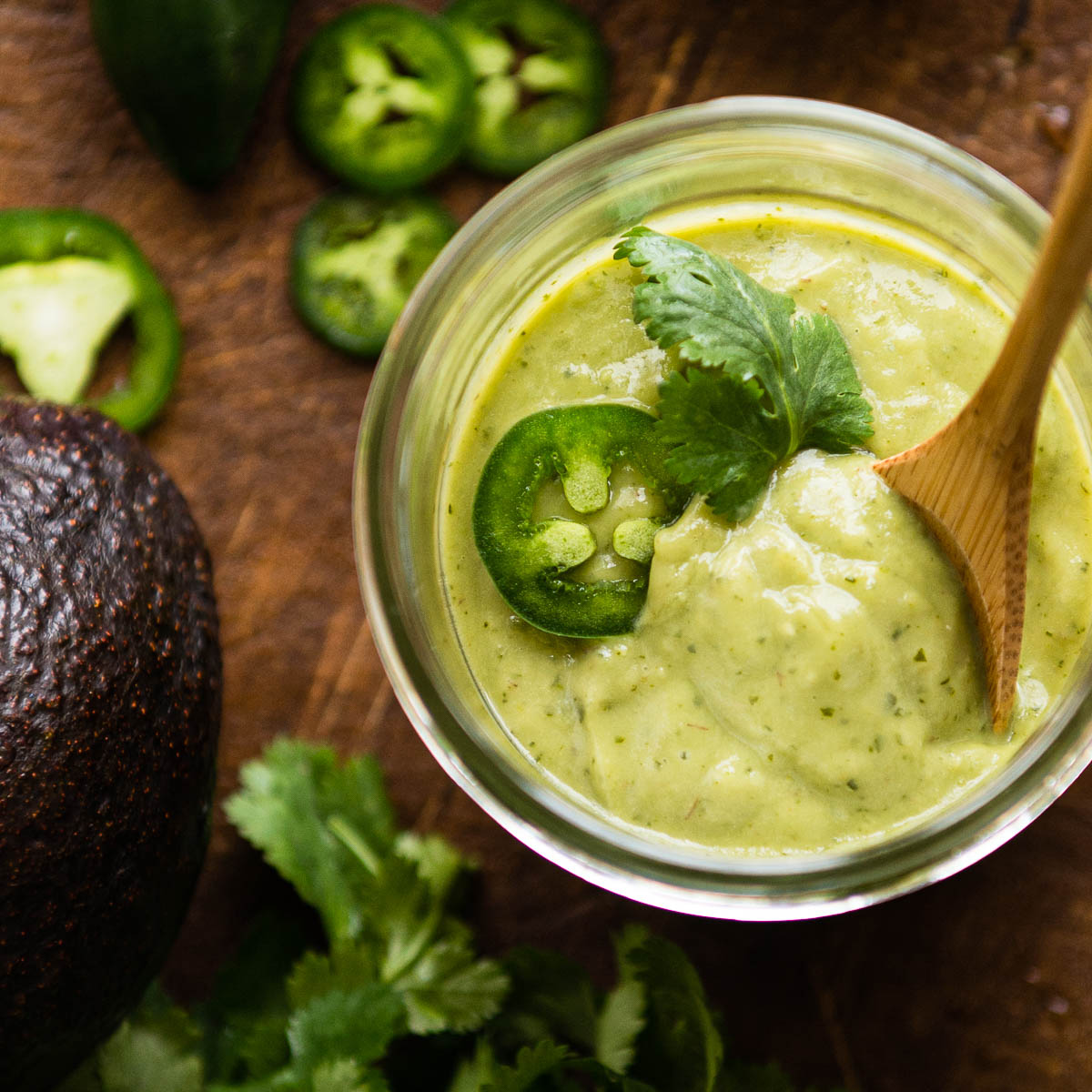 vegan avocado crema in a glass jar topped with cilantro, sliced jalapeño and a wooden spoon.