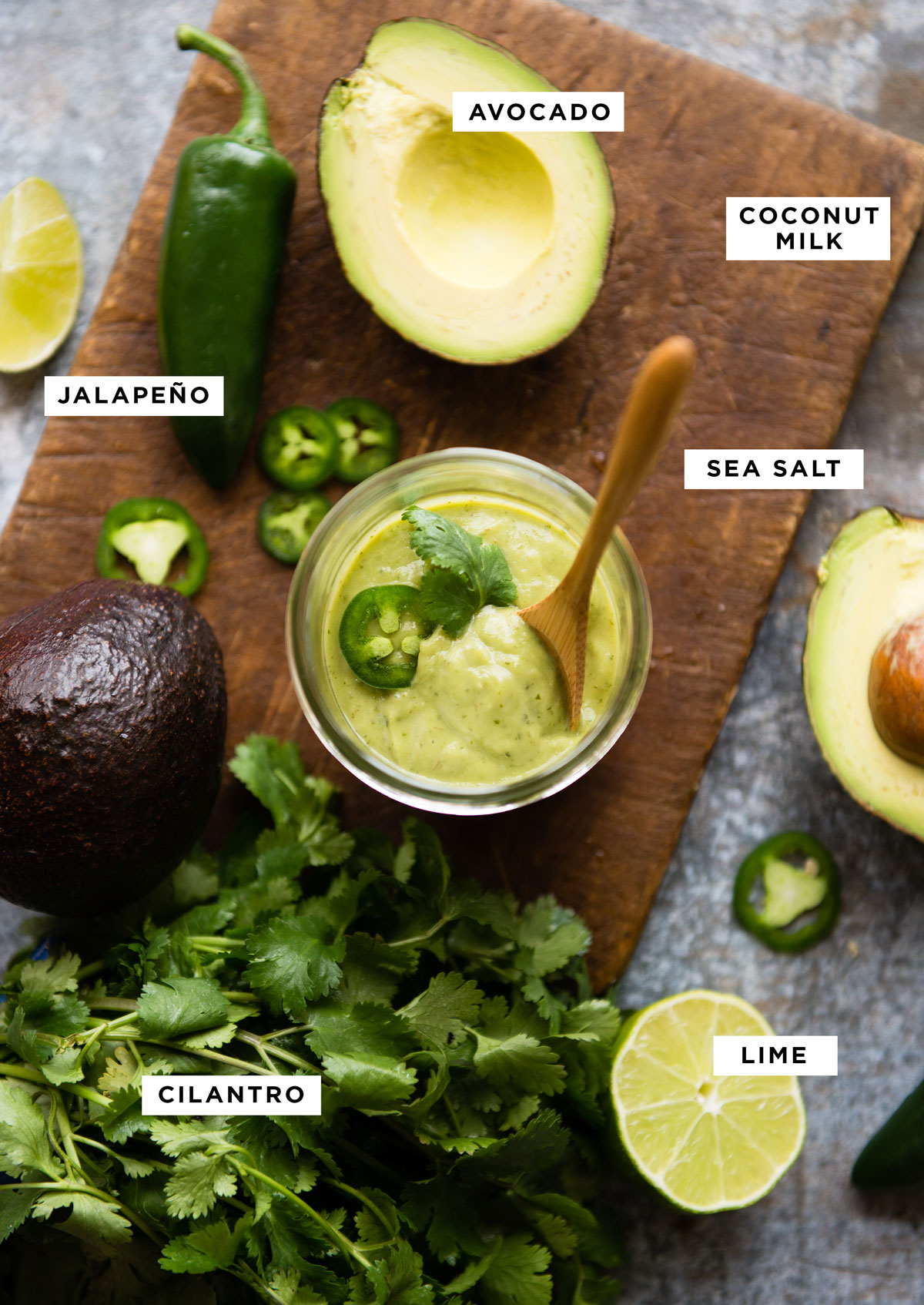 Labeled ingredients for avocado crema recipe including avocado, coconut milk, jalapeño, sea salt, lime and cilantro on a wood-grain cutting board.