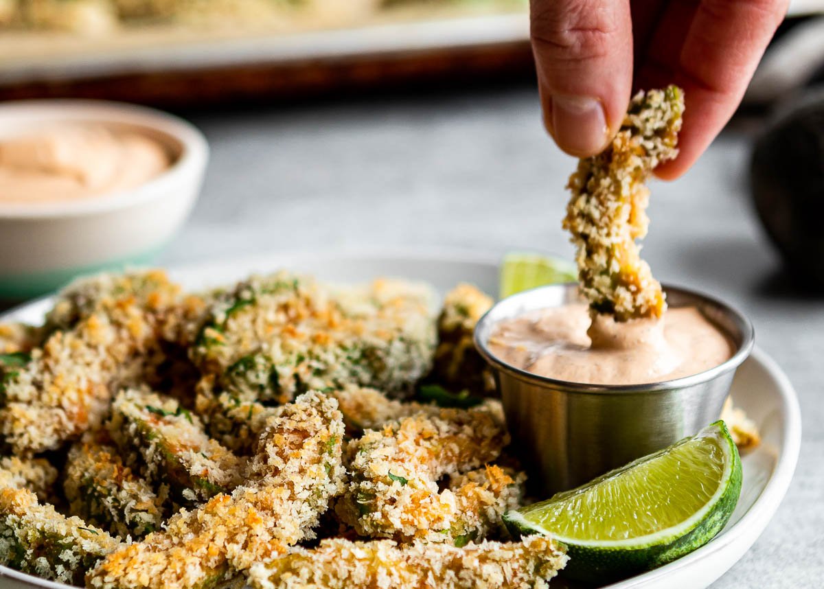 dipping a plate of avocado fries in chipotle ranch dipping sauce with lime wedges.