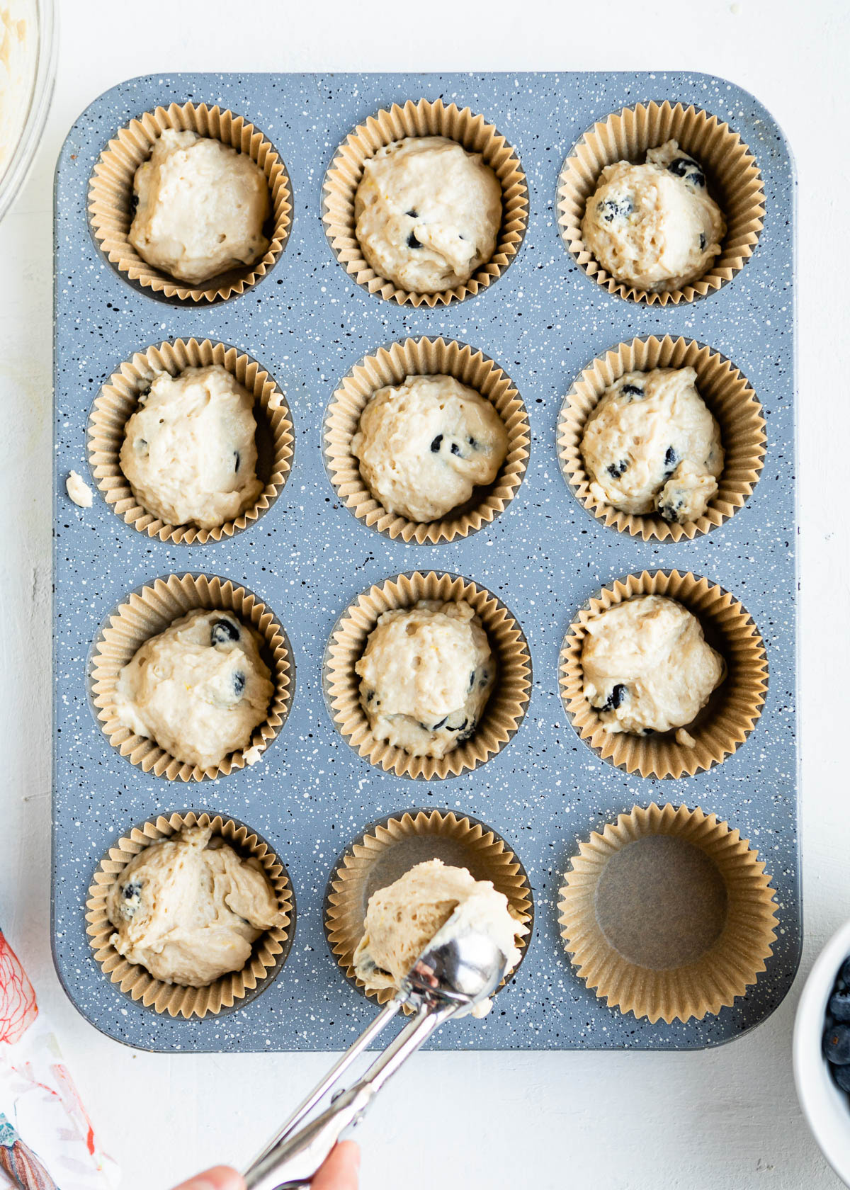 using a metal scoop to put vegan blueberry muffin batter into lined muffin tins.