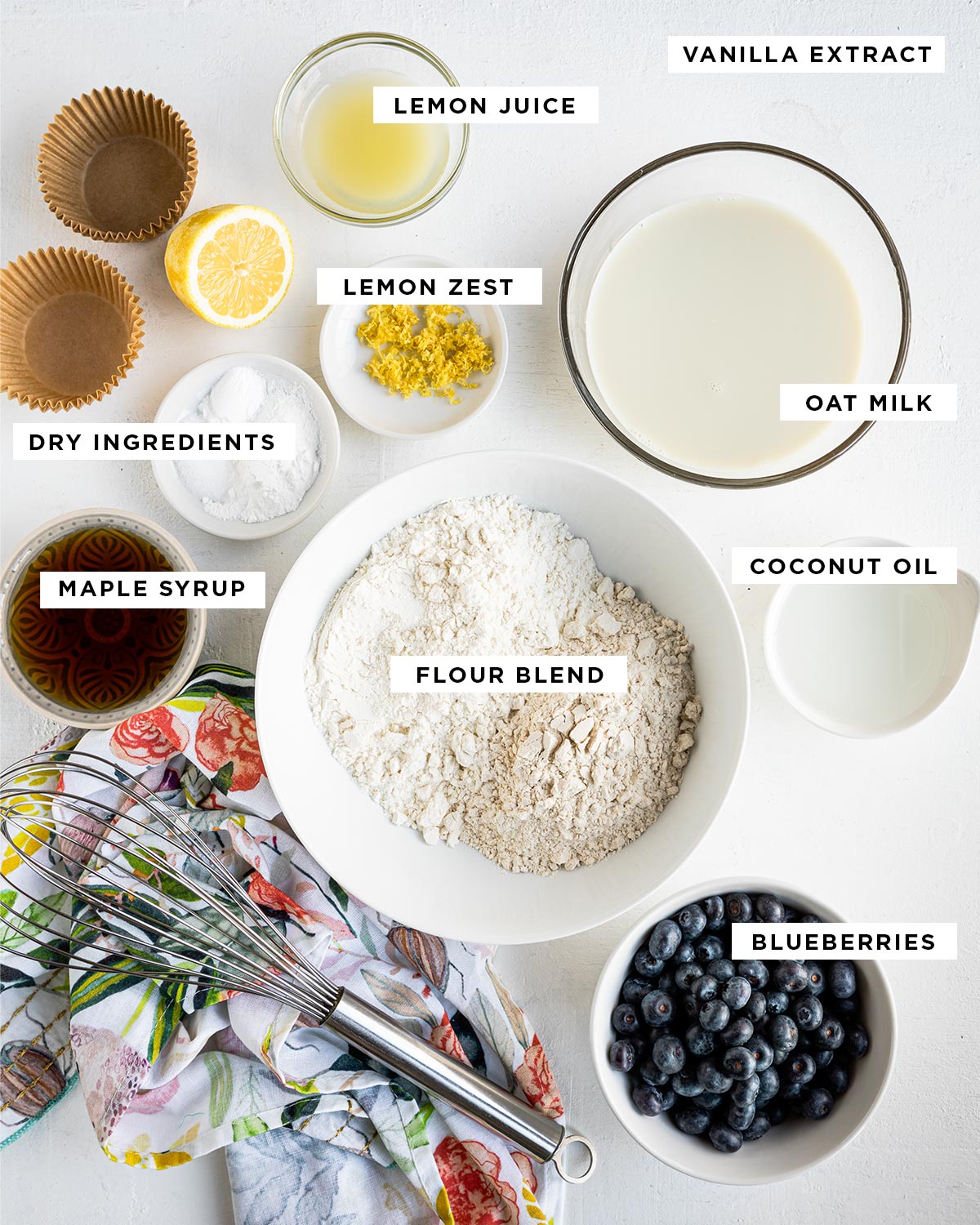 labeled ingredients for vegan muffins including lemon juice, vanilla extract, lemon zest, oat milk, dry ingredients, maple syrup, coconut oil, flour blend and blueberries.
