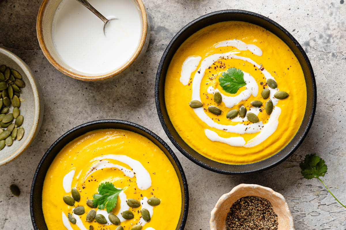 Two black bowls filled with Carrot ginger Soup topped with swirls of cashew cream, fresh cilantro, and pepitas.