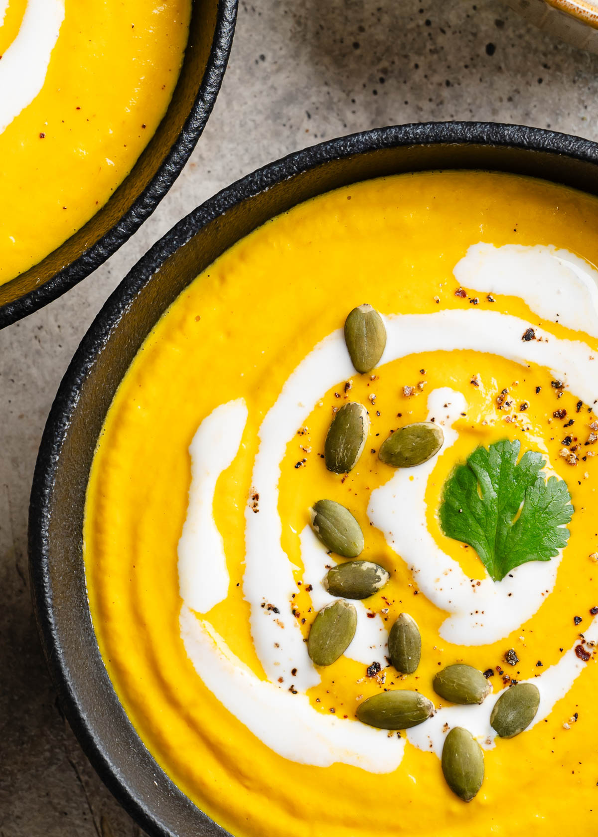 A black bowl filled with Carrot ginger Soup topped with swirls of cashew cream, fresh cilantro, and pepitas.