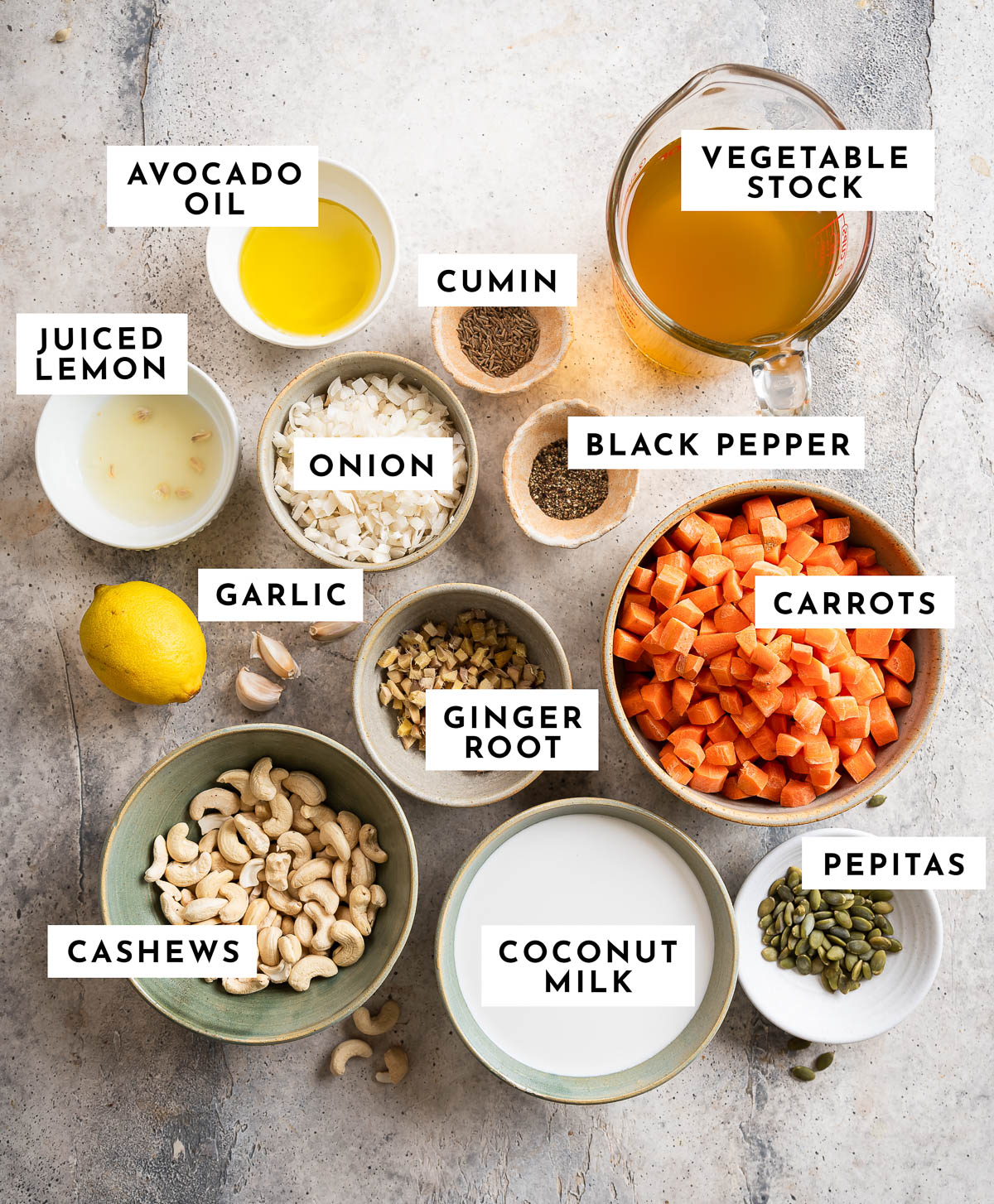 Labeled ingredients for making coconut ginger soup, Including avocado oil, onions, ginger root, garlic cloves, cumin, carrots, vegetable stock, coconut milk, cashews, lemon juice, sea salt, and black pepper.