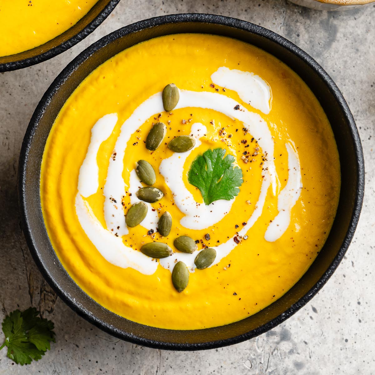 A black bowl filled with Carrot ginger Soup topped with swirls of cashew cream, fresh cilantro, and pepitas.