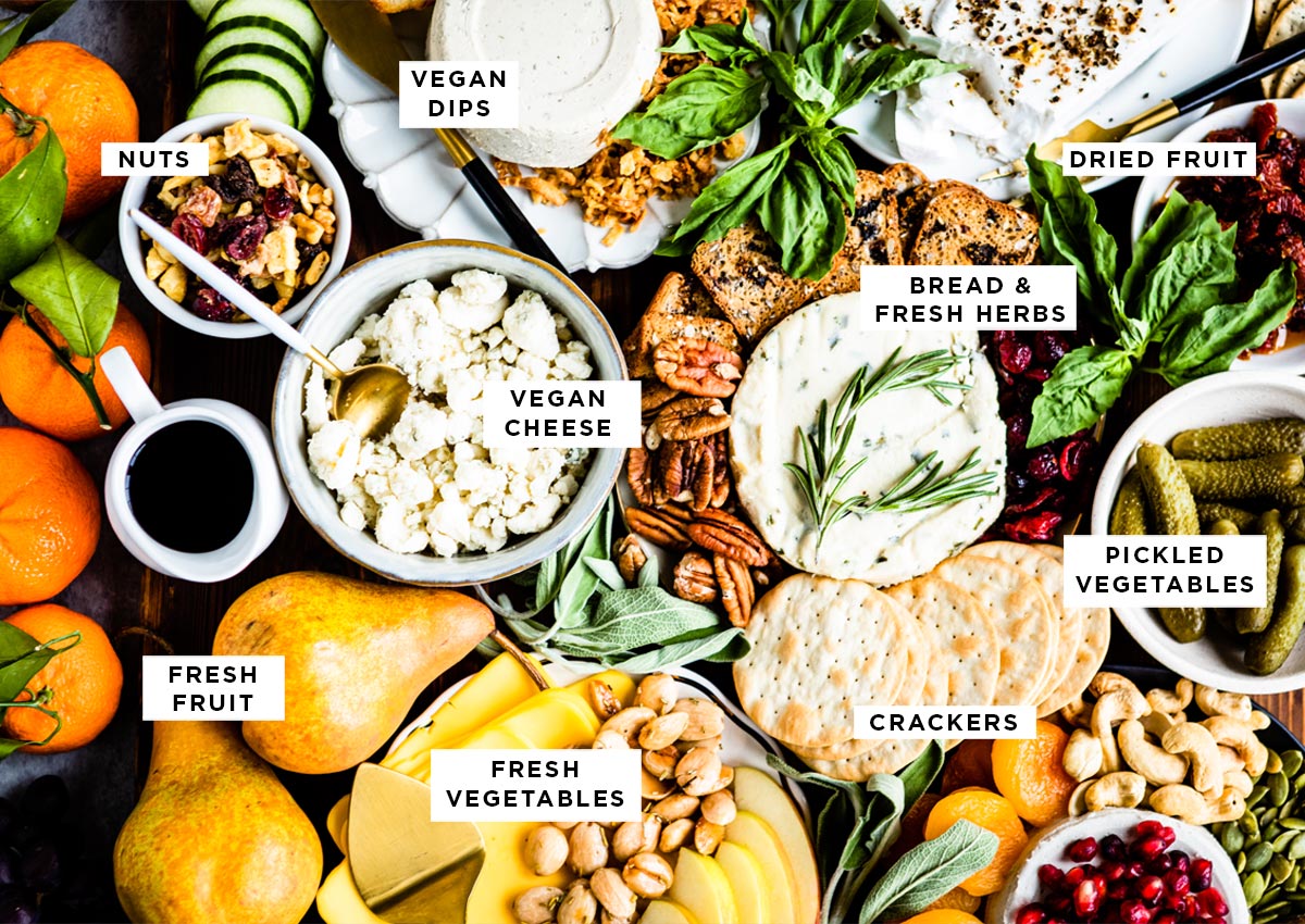 ingredients for a plant-based charcuterie board including vegan dips, nuts, dried fruit, bread and fresh herbs, vegan cheese, fresh fruit, fresh vegetables, crackers and pickled vegetables.