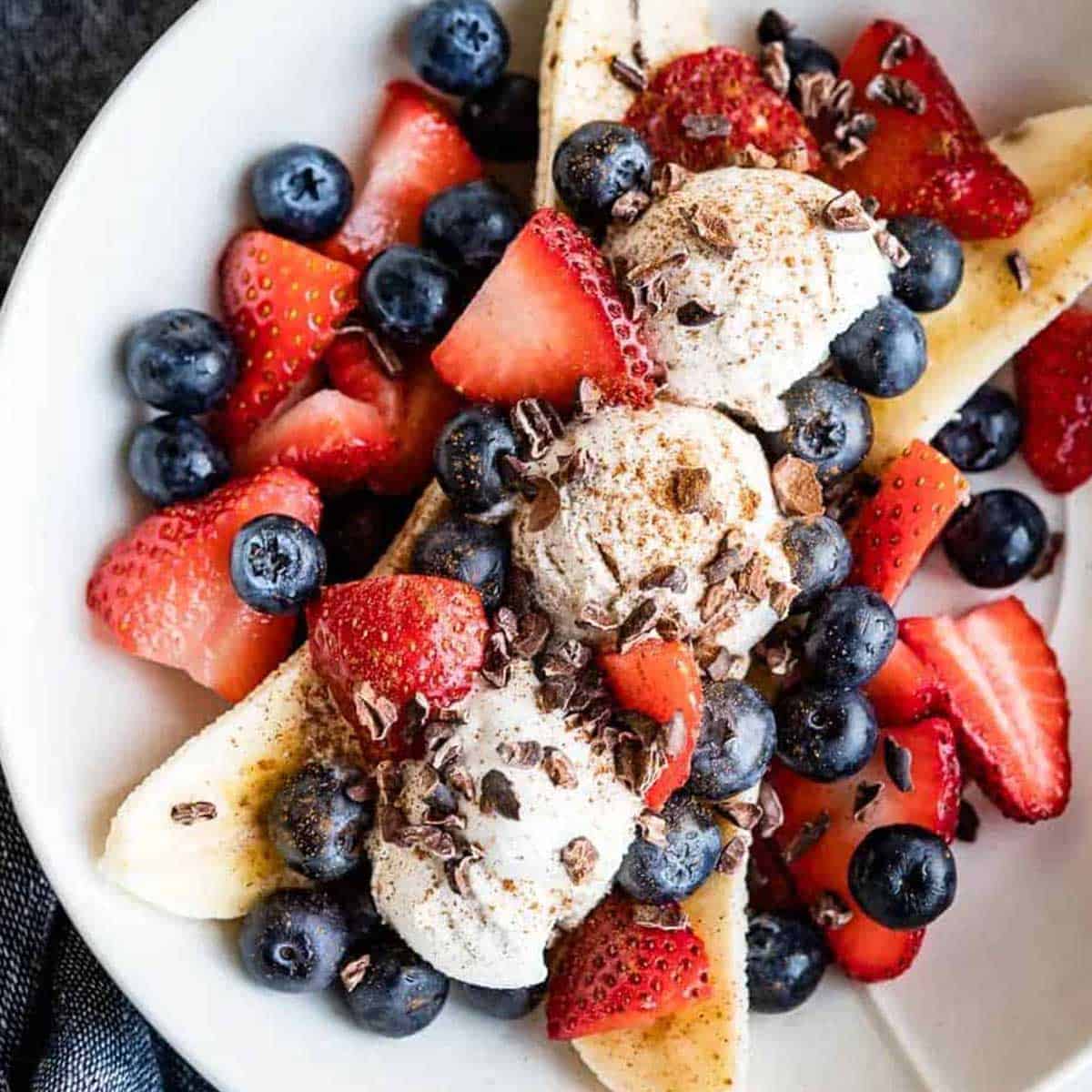 banana split with 3 scoops of ice cream in between a banana sliced in half lengthwise and topped with fresh strawberries, blueberries and cacao nibs.