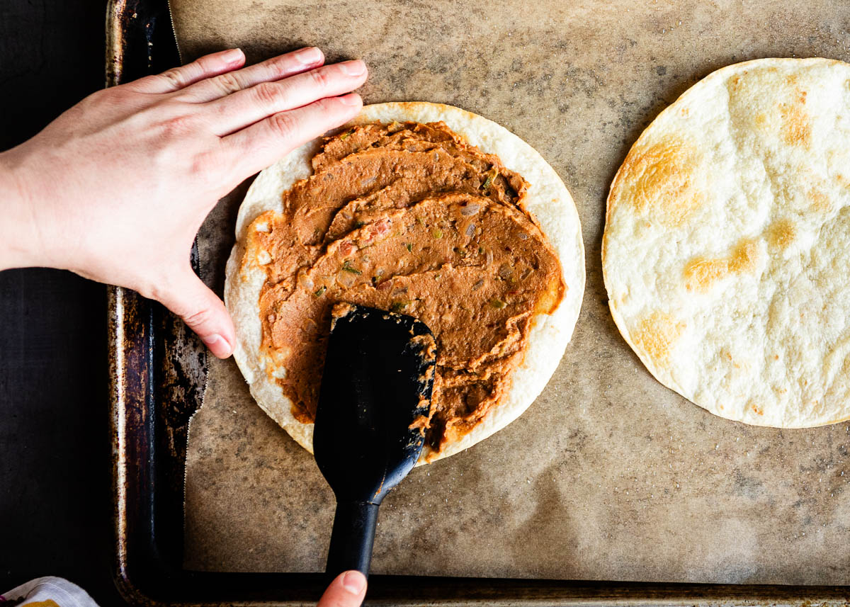 spreading refried beans with a black spatula onto crispy tortilla shells.