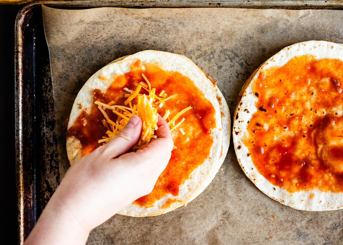 sprinkling vegan cheddar cheese on top of hot sauce on tortilla shells.