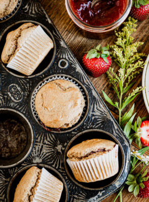 Antique black muffin tin with baked vegan muffins in white wrappers, next to strawberry jam.