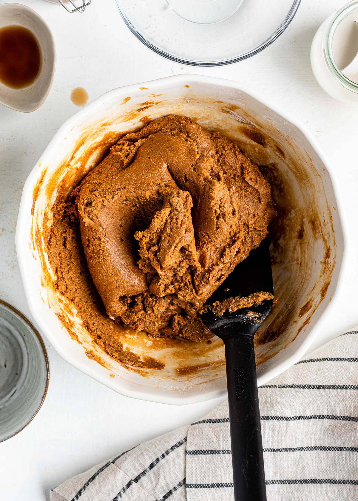 caramel colored cookie dough getting mixed in a white mixing bowl by a black spatula.