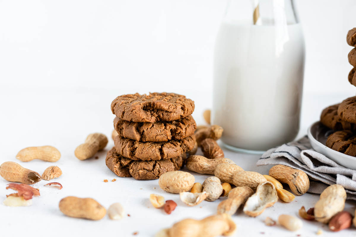 stack of eggless peanut butter cookies on a white counter top next to a glass jar of oat milk, surrounded by shelled peanuts.