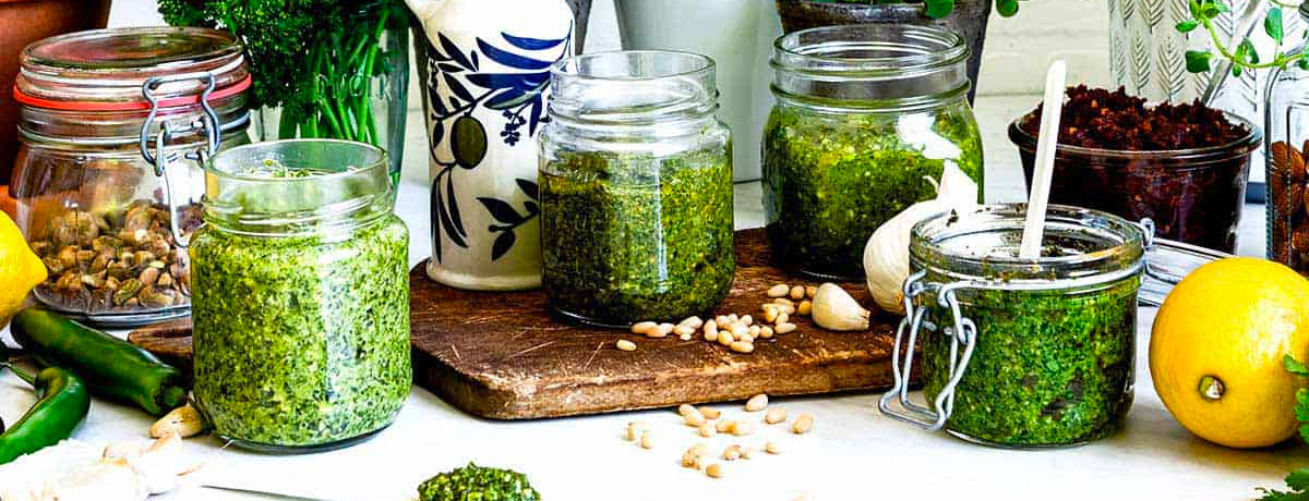 four jars of pesto on a table with varieties of herbs scattered around