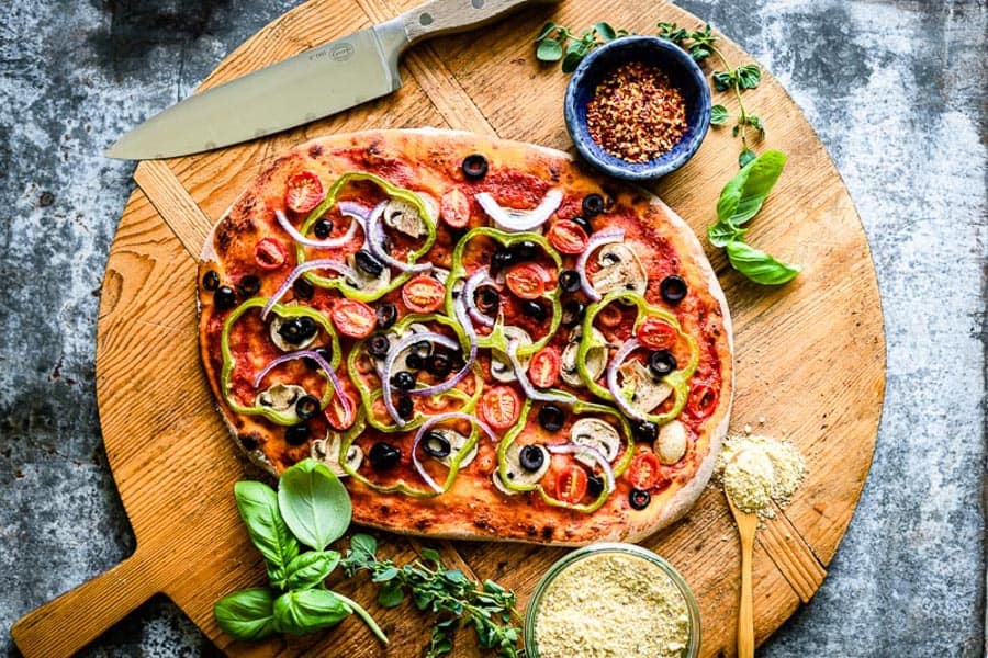 vegan pizza recipe with fresh toppings surrounding it on a wooden tray.