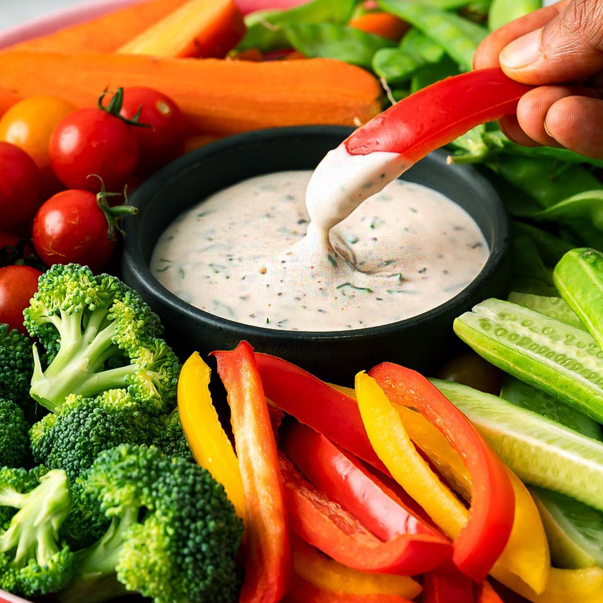 Dark bowl filled with vegan ranch dip, surrounded by colorful assortment of fresh vegetables.