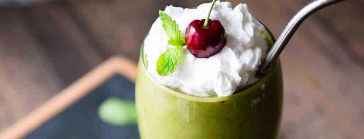 green smoothie in a fancy glass with whipped cream on top and a dark red cherry