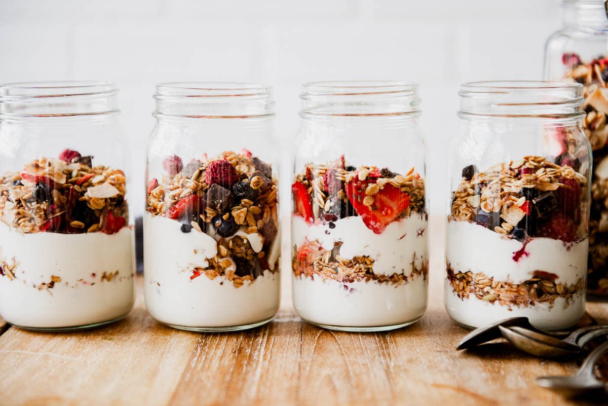 4 glass containers of vegan yogurt layered with granola and fresh berries next to a pile of spoons.
