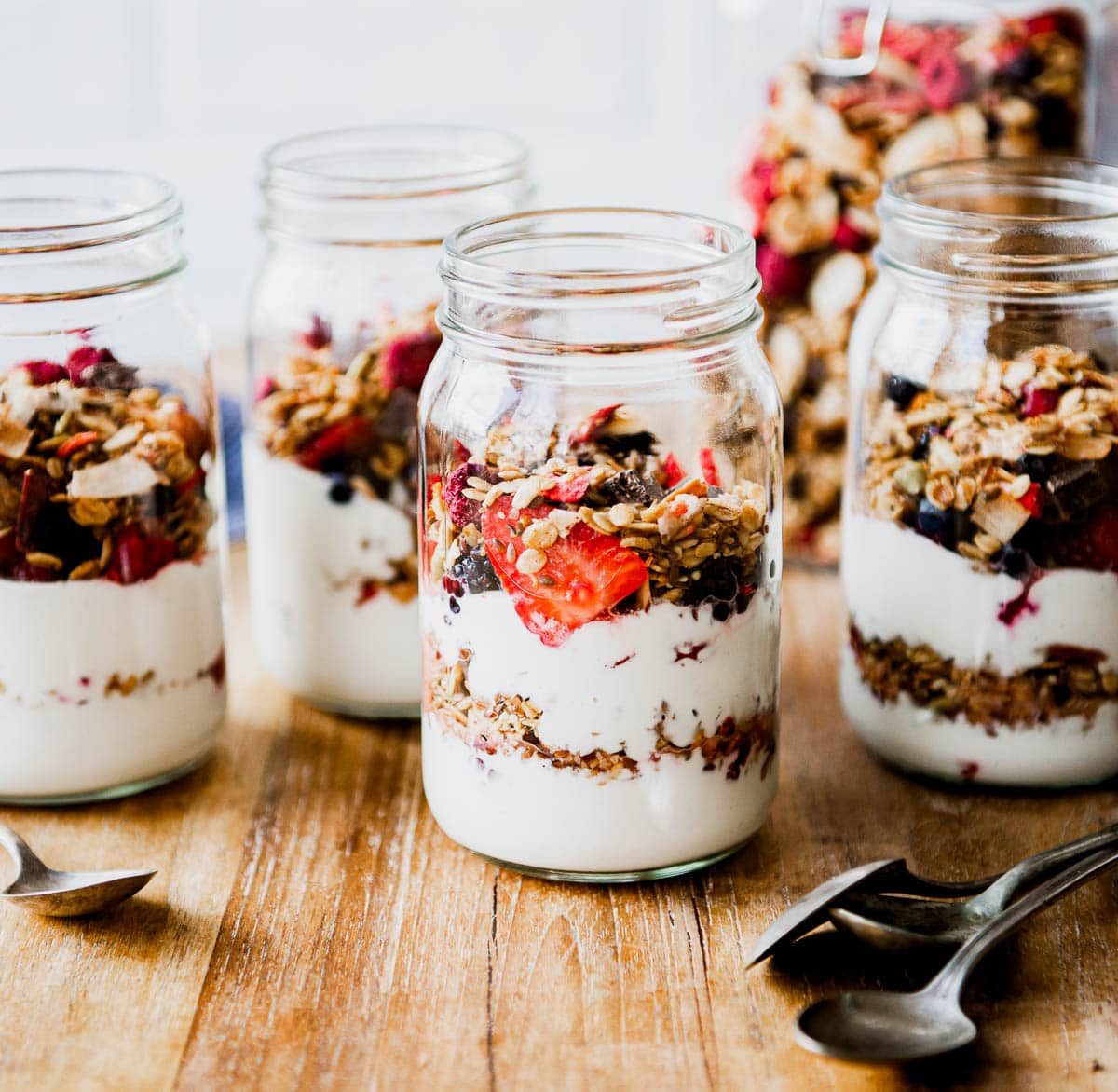 several glass jars of plant based yogurt mixed with granola and fresh berries on a wooden table top, next to a pile of spoons.
