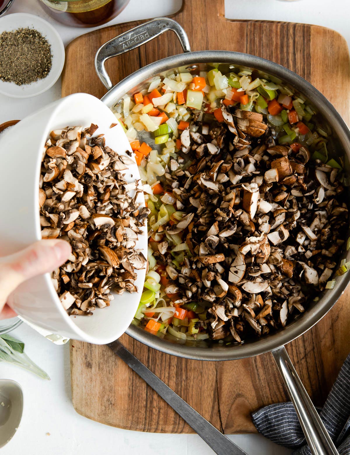 Person pouring chopped mushrooms into a pan with vegetables.