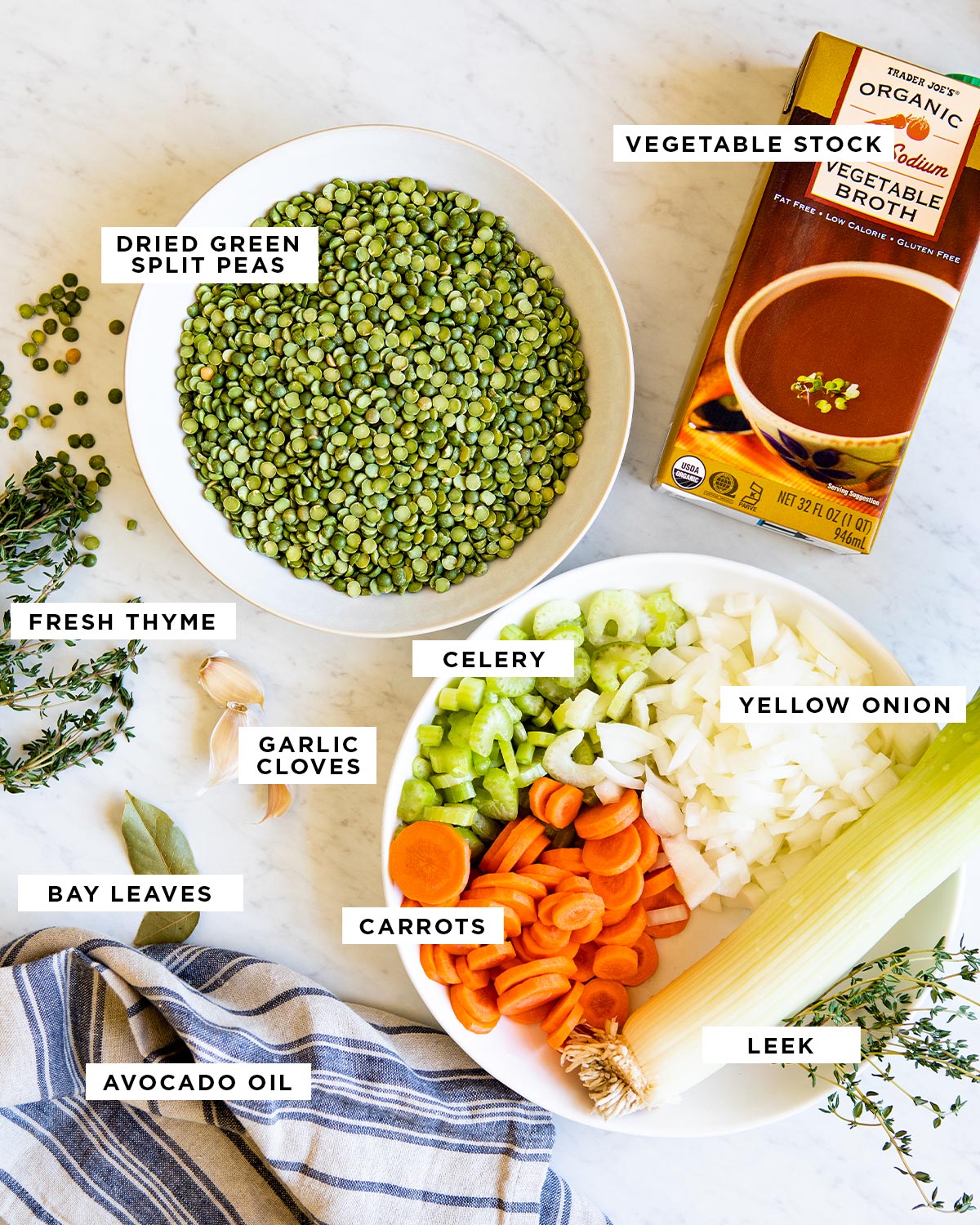 Ingredients for vegetarian split pea soup on marble countertop including vegetable stock, dried green split peas, fresh thyme, celery, yellow onion, garlic cloves, bay leaves, carrots, leek and avocado oil.