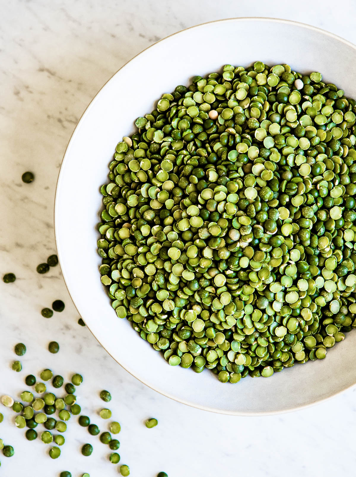 Large white bowl of uncooked green split peas on marble countertop.