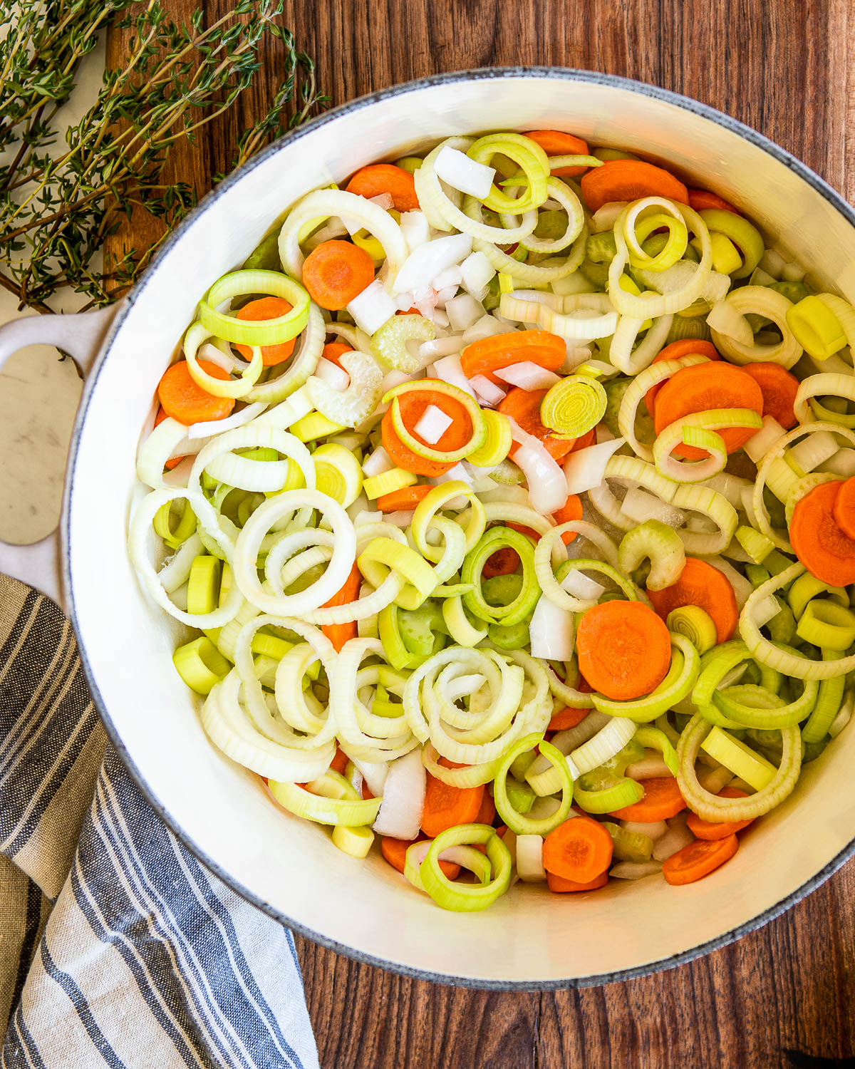 White stock pot with onions, carrots, celery and leeks then another white stock pot getting filled with vegetable stock.