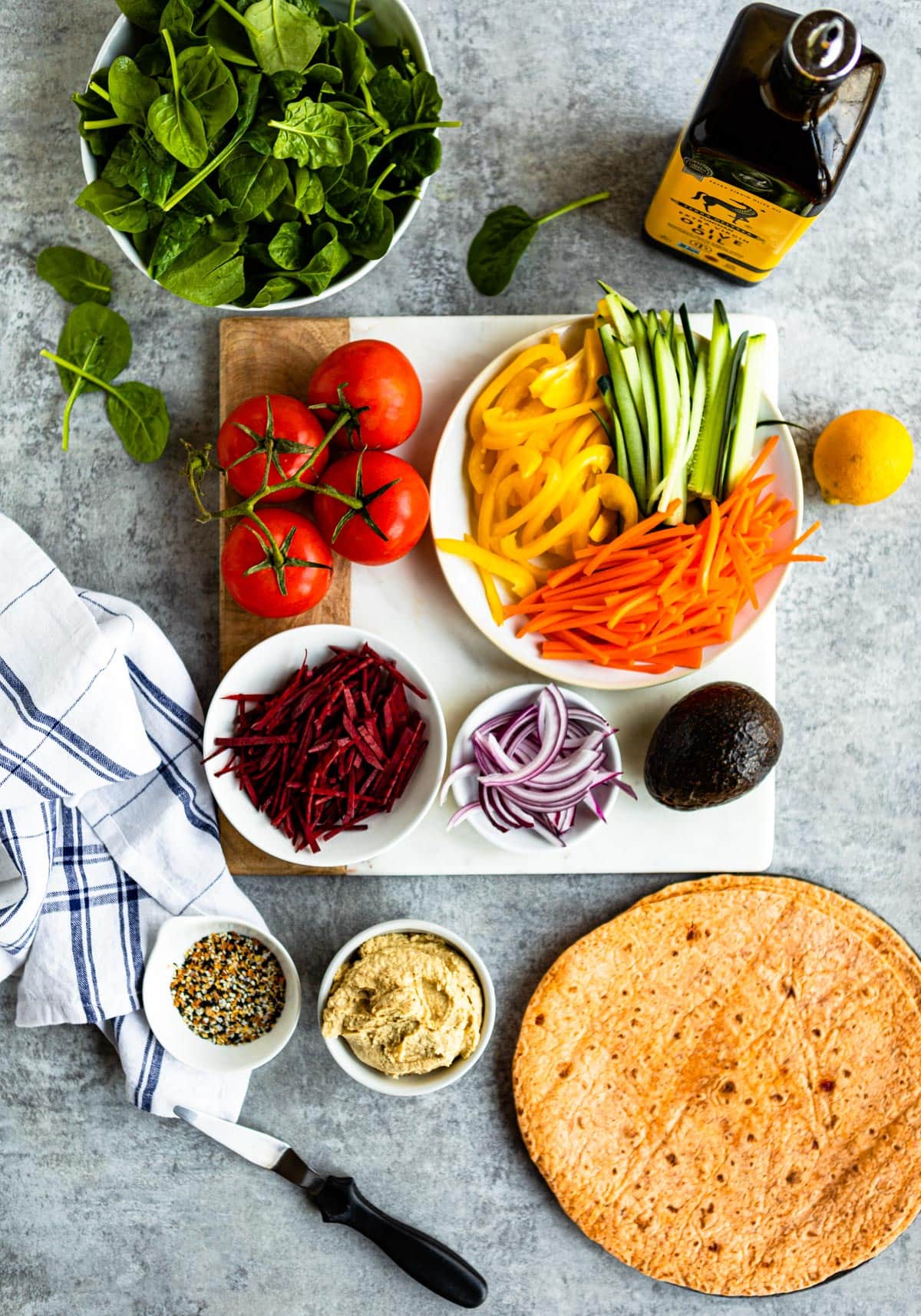 Veggie wrap ingredients for a healthy lunch, including a rainbow of vegetables, everything bagel seasoning, hummus, and some sun-dried tomato tortilla shells.