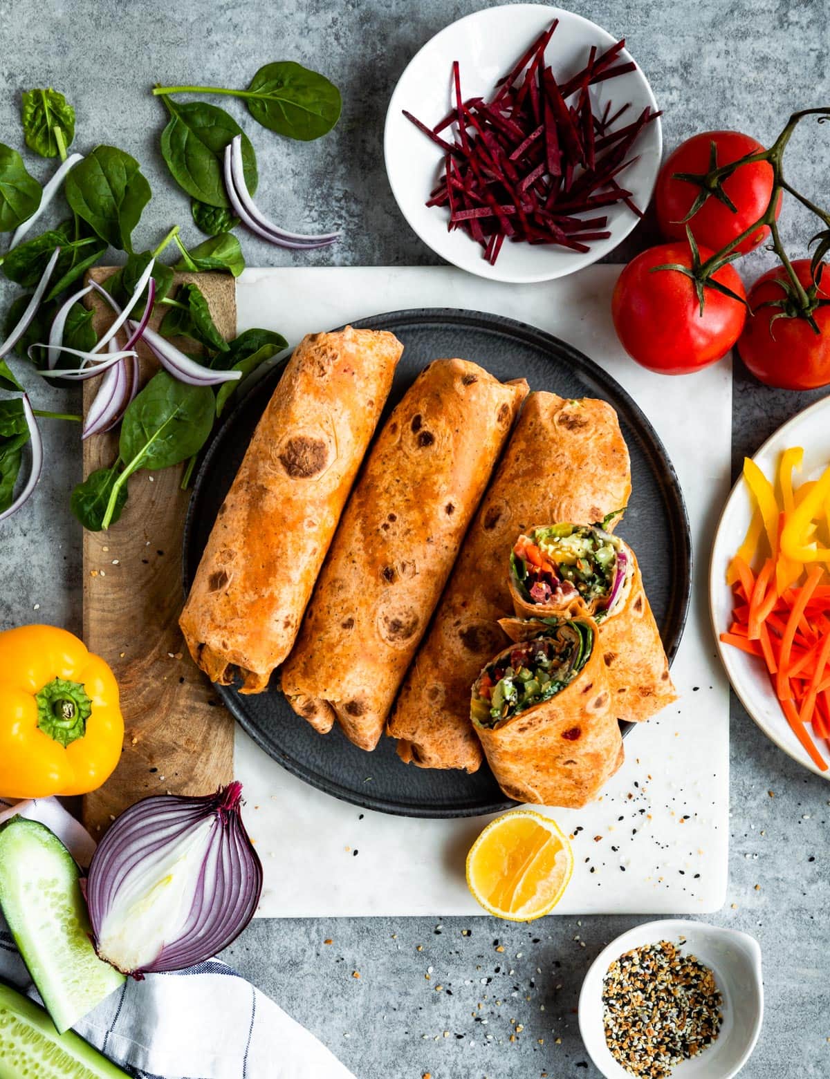 4 hummus wraps on a gray plate surrounded by fresh vegetables.