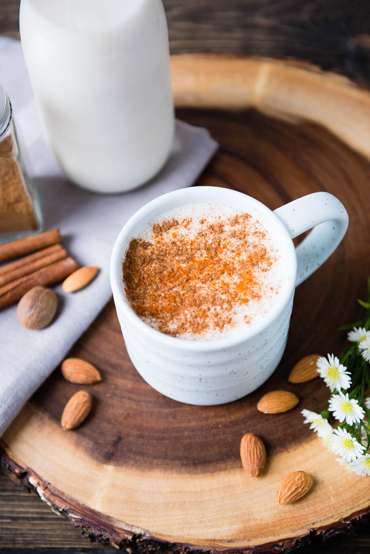 image of warm almond milk topped with spices in a white ceramic mug surrounded by raw almonds, nutmeg and cinnamon