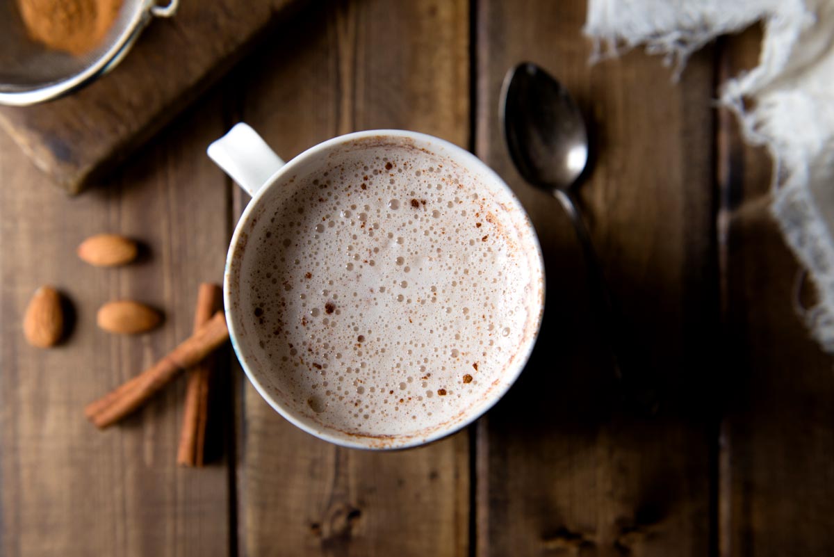 A mug of warm, frothy almond milk topped with a sprinkle of cinnamon and cayenne pepper. Cinnamon sticks, almonds, and ground ginger are in the background. 