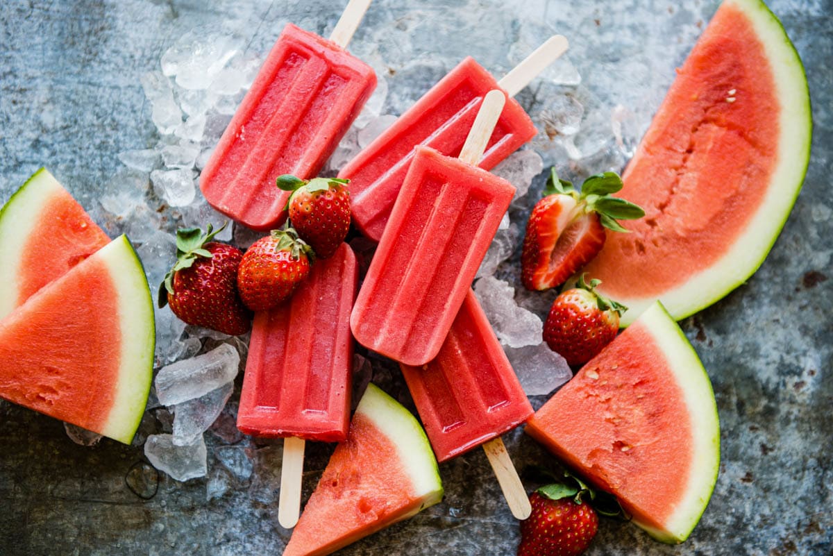 watermelon popsicles sitting on ice cubes with whole strawberries and watermelon slices around them.