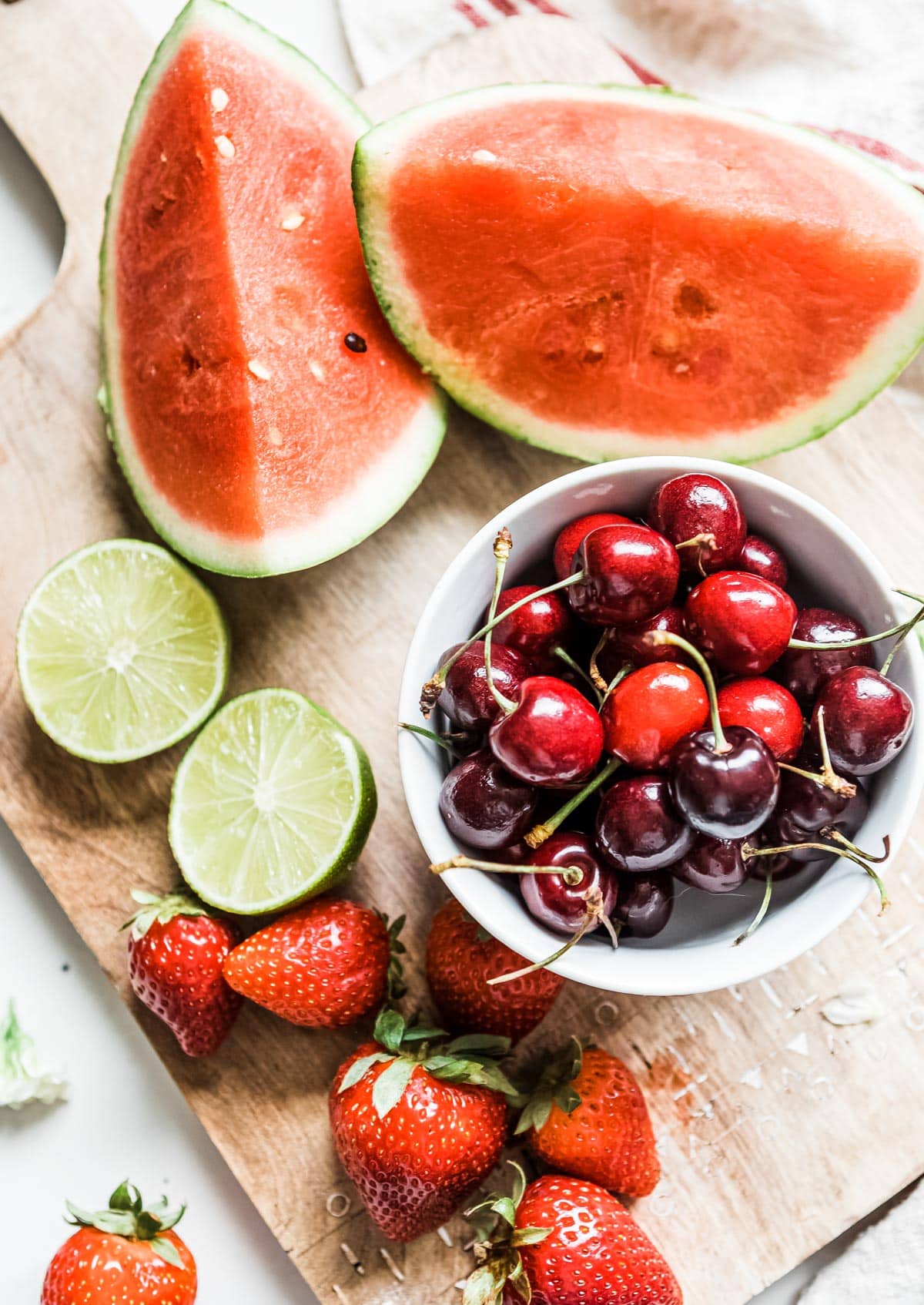 ingredients for a delicious summer smoothie on a wooden cutting board including watermelon, lime, cherries and strawberries
