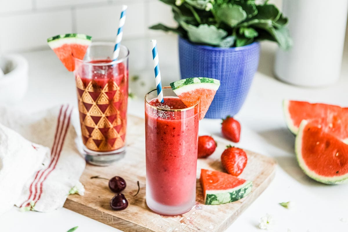 2 glasses of red smoothie surrounded by fresh cherries, strawberries and watermelons.