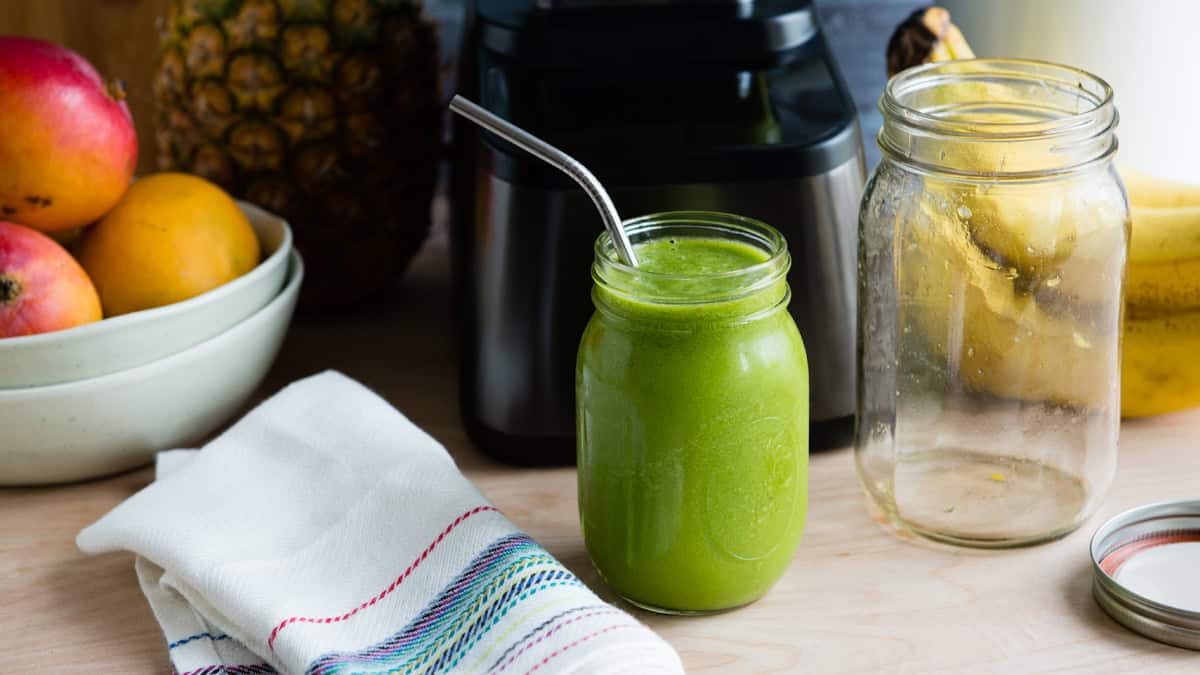 Healthy green smoothie in a glass jar with fresh fruits and blender in background.