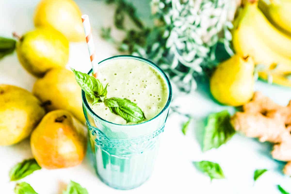 A glass of the best smoothie for acid reflux, with a straw and topped with fresh basil. Fresh ingredients are blurred in the background.