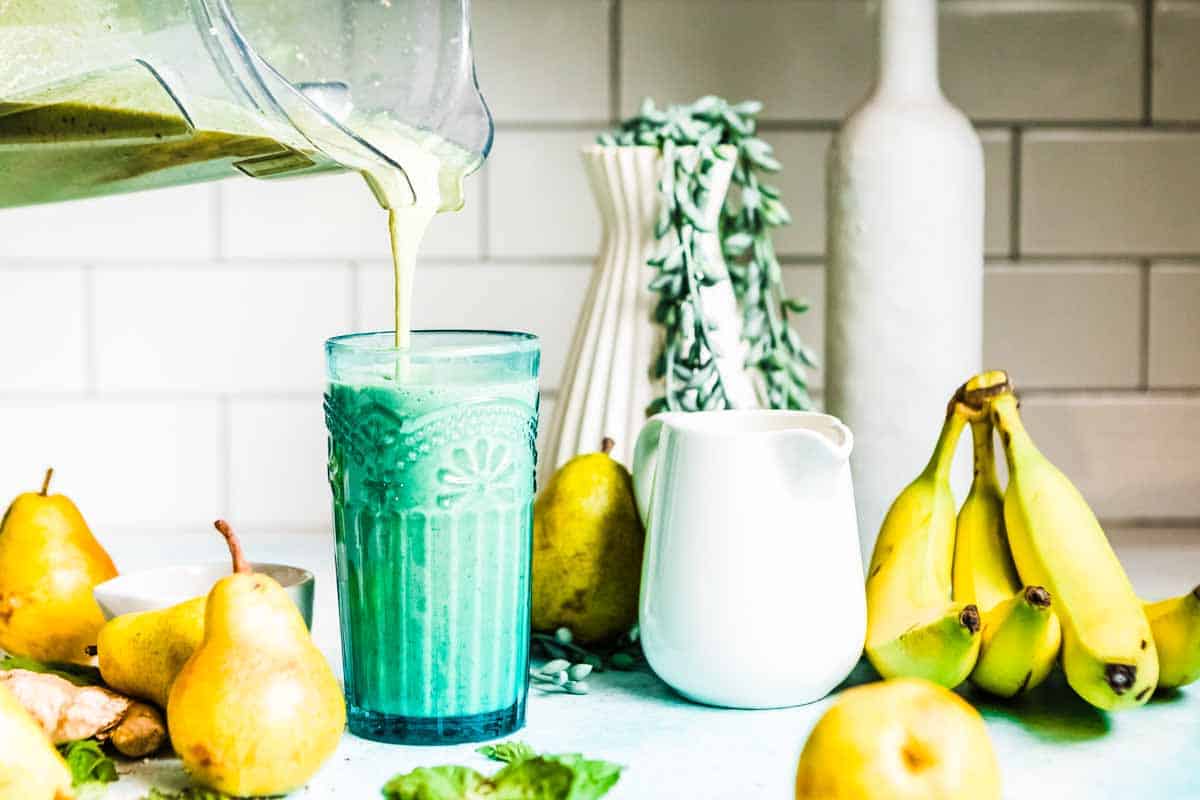 Fruit smoothie for acid reflux pouring into a glass, on a countertop, surrounded by pears and bananas.